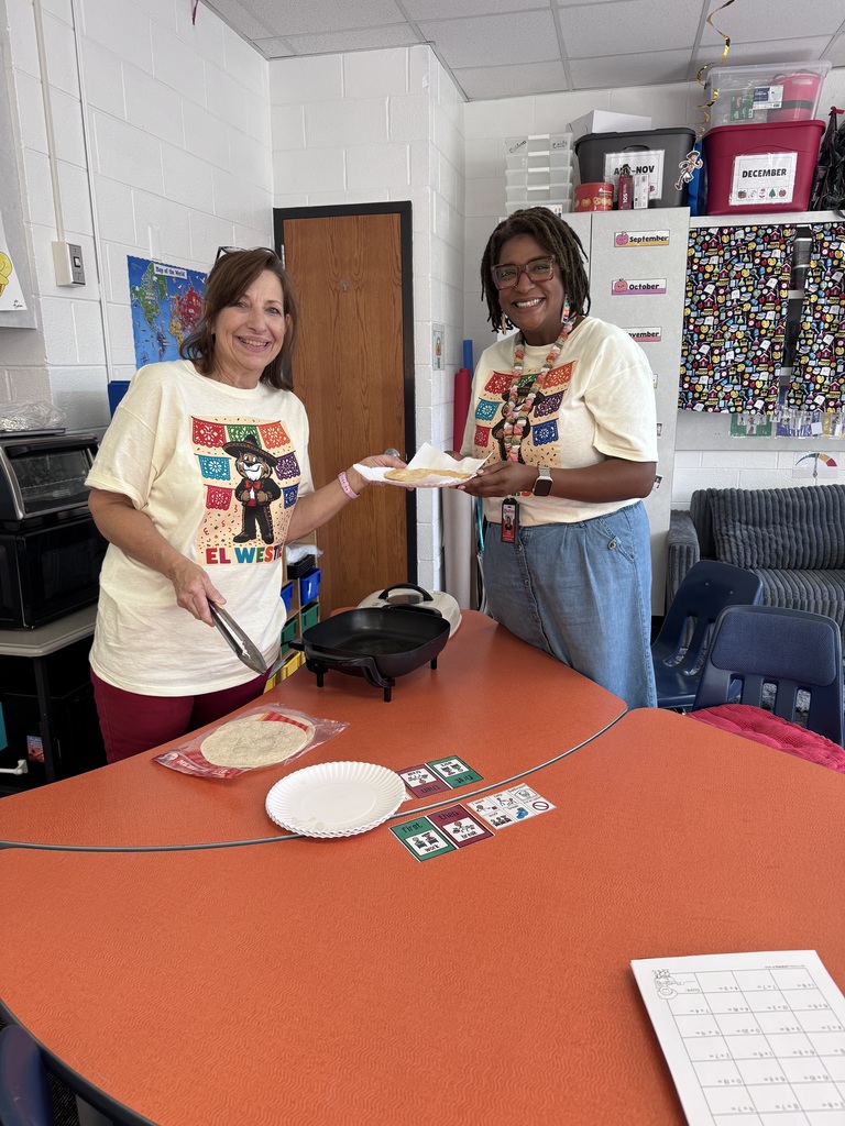 Two educators smile in a classroom while holding up a paper plate with freshly made fry bread over an orange table.    Foreground & Educators:  The educator on the left has brown hair and glasses on her head, wearing a cream "El Westie" graphic t-shirt and dark pink pants while holding metal tongs above an electric skillet.    The educator on the right has dark locs and glasses, wearing a matching "El Westie" shirt and denim skirt, smiling as she holds the plate of fry bread.    The orange table displays paper plates, a package of tortillas, educational visual schedules, and a math worksheet.    Background: An electric skillet sits on the table in front of classroom storage cabinets, labeled bins, and a world map on the white cinderblock wall.