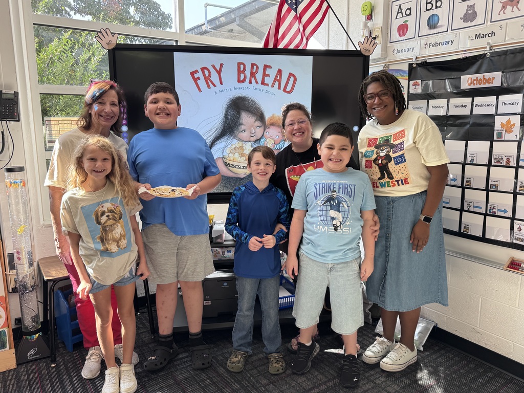 Six people standing together in a classroom pose for a group photo in front of a digital screen displaying the book cover for Fry Bread: A Native American Family Story.    Front Row (Left to Right):  A young girl with long, wavy blonde hair wearing a graphic t-shirt featuring a Shih Tzu dog, denim shorts, and white sneakers.    A young boy in a long-sleeved blue pattern shirt and jeans smiling while wearing green patterned clogs.    A young boy in a light blue graphic t-shirt that says "STRIKE FAST / MESSI", light denim shorts, and black sneakers smiling broadly.    Back Row (Left to Right):  An adult woman with colorful highlights in her hair wearing a light shirt, pink pants, and white sneakers.    A taller boy in a bright blue t-shirt and grey shorts holding a paper plate with a piece of decorated fry bread.    An adult woman wearing glasses and a dark t-shirt with a red design standing behind the boys.    An adult woman with glasses and curly dark hair wearing a yellow t-shirt reading "EL WESTIE", a long blue denim skirt, and white sneakers.    Background: An American flag hangs above the screen. To the right, an October calendar and alphabet posters hang on the wall, with a decorative water tube light on the left.