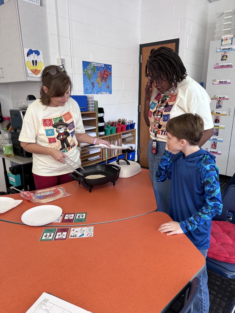 Two educators and a young student gather around a kidney-shaped orange table in a classroom to prepare food together.    Left: An adult woman with shoulder-length brown hair and glasses resting on her head wears a cream-colored "El Westie" t-shirt and magenta pants. She uses metal tongs to lift fry bread out of an electric skillet.    Right: An adult woman with dark locs and glasses wears a matching "El Westie" t-shirt and blue jeans, looking down attentively with one hand near her face. Beside her, a young boy in a blue long-sleeved camo-patterned shirt leans forward, watching the cooking process closely.    Tabletop & Environment: The orange table holds paper plates, a package of tortillas, a white appliance lid, and visual schedule cards. In the background, classroom storage shelves, a world map, and a birthday chart line the white cinderblock wall.