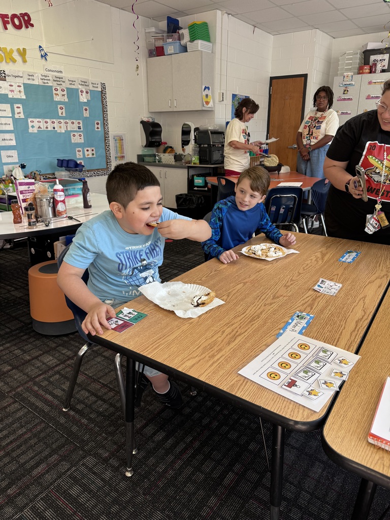 Two students sit at a light wooden table in a classroom, eating decorated fry bread.    Foreground:  A young boy on the left in a light blue "STRIKE FAST / MESSI" t-shirt takes a bite of his fry bread.    Beside him, a boy in a long-sleeved blue camo shirt smiles while watching his classmate, with a plate of topped fry bread in front of him.    On the table lie communication cards, name tags, and an educational feedback chart.    Background: An adult woman in a black t-shirt stands to the right holding a phone, while two other educators work near a cooking station in the background beside classroom storage cabinets and a phonics board.