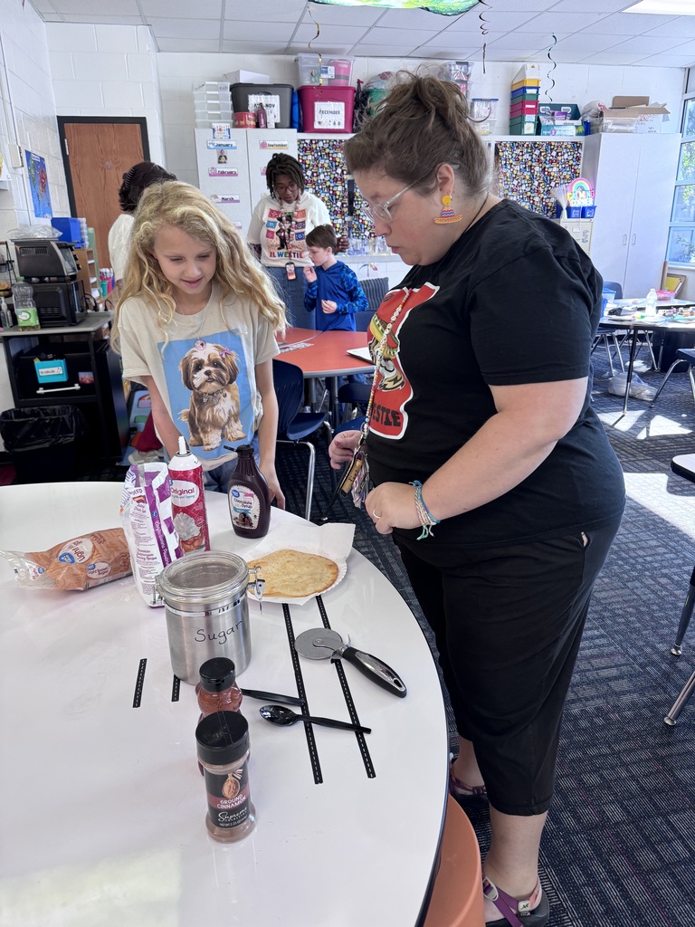 A student and an educator stand at a round white table in a classroom, preparing sweet toppings for fresh fry bread.    Foreground:  A young girl with long, wavy blonde hair wearing a shirt printed with a Shih Tzu looks down with a smile.    An adult woman wearing glasses, decorative earrings, and a black t-shirt leans over the table to assist her.    The table holds a piece of fry bread on a paper plate alongside chocolate syrup, whipped cream, powdered sugar, brown sugar, a sugar canister labeled "Sugar", ground cinnamon, plastic utensils, and a pizza cutter.    Background: In the background, another educator and student stand near an orange table, with classroom storage cabinets and a "Calming Corner" area visible on the right.