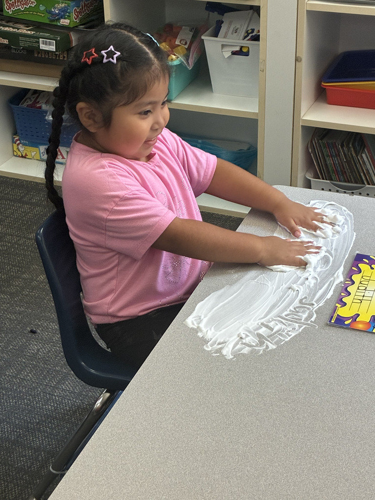 A young girl with braided hair and star clips sits at a classroom table, smiling as she spreads white shaving cream across the tabletop with both hands. Next to her is a colorful nameplate that reads "Judith".