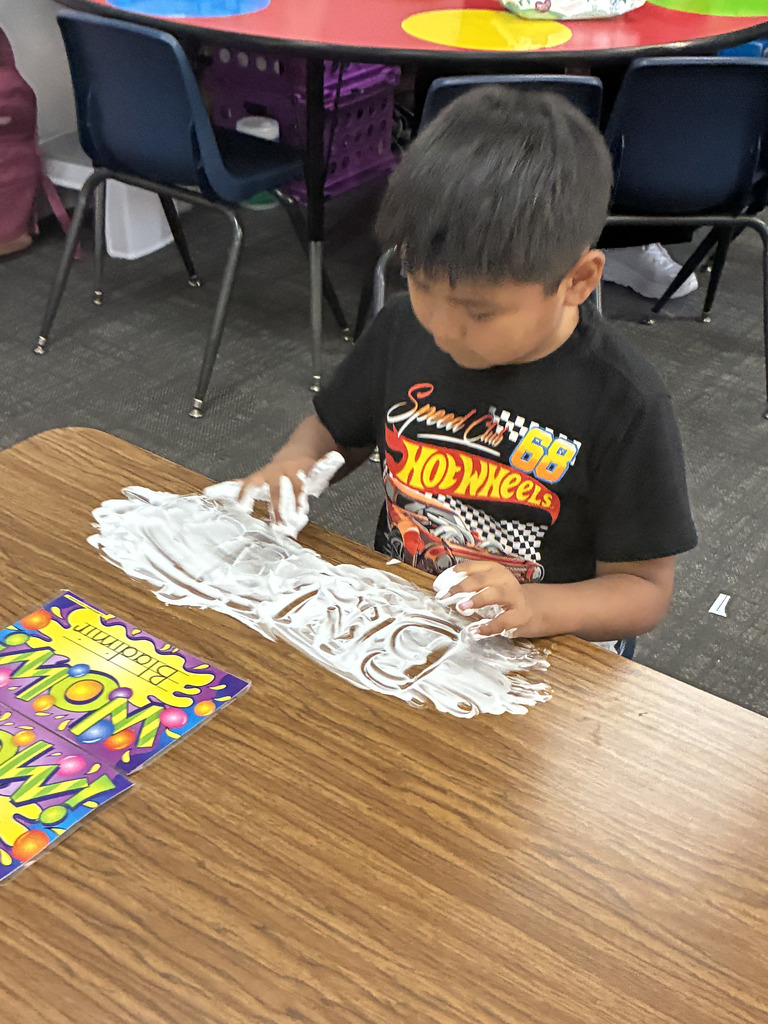 A young boy in a black Hot Wheels graphic t-shirt leans over a wooden desk, using his fingers to trace letters into shaving cream spread on the desk surface. A bright name tag labeled "Bladimir" lies next to his workspace.