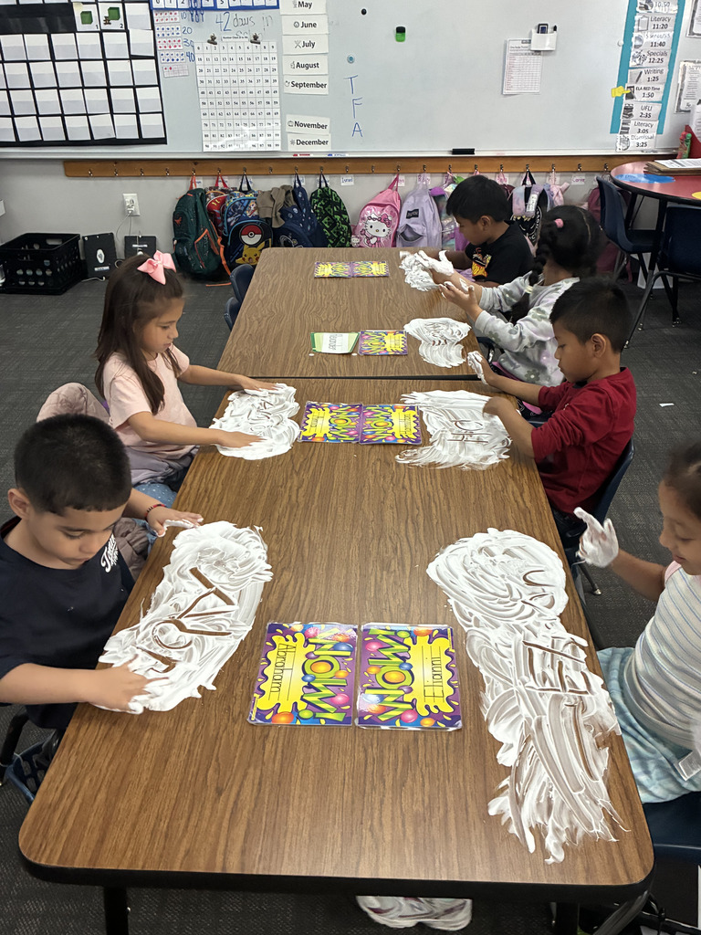 High-angle shot of a group of elementary students sitting along connected classroom tables. The students are actively writing their names with their fingers into white shaving cream spread across the wooden surface. Classroom calendars and hanging backpacks are visible in the background.
