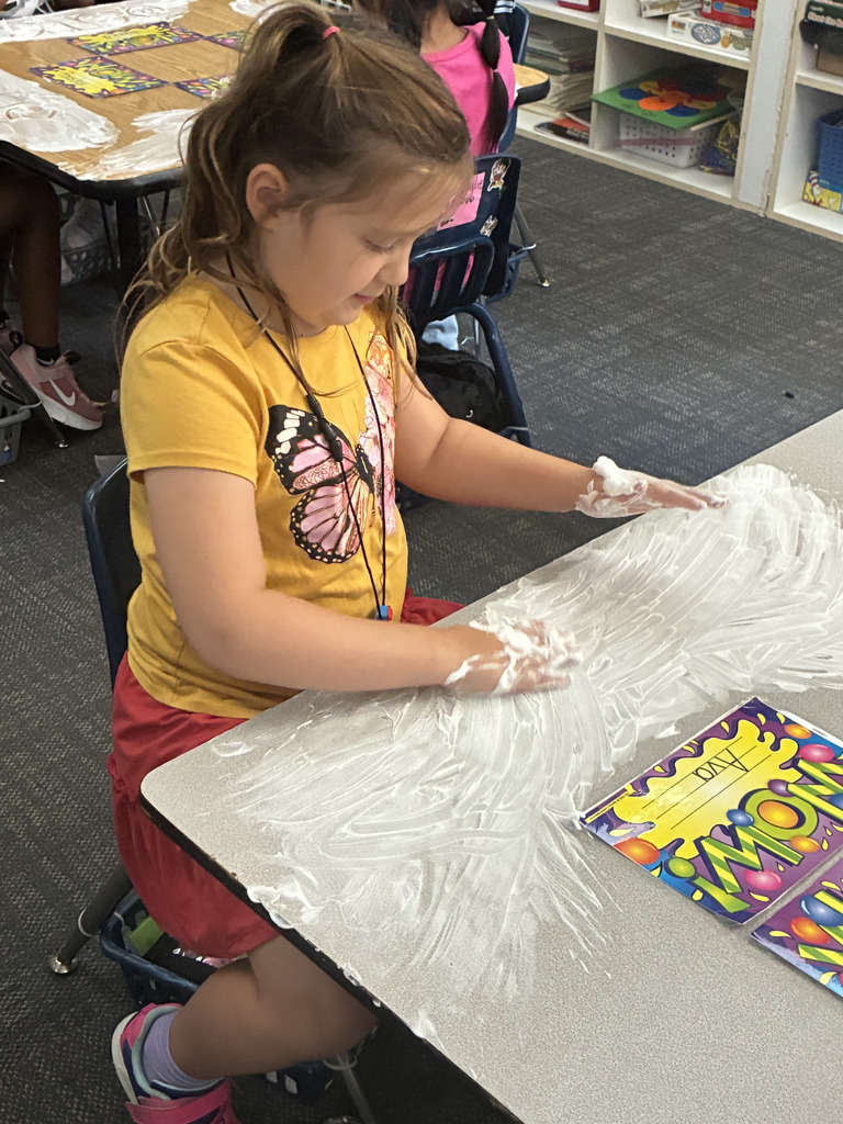 A young girl wearing a yellow butterfly shirt sits at a gray table, using both hands covered in white shaving cream to smooth it out across the desk surface. A yellow name tag that reads "Ava" sits beside her work area.