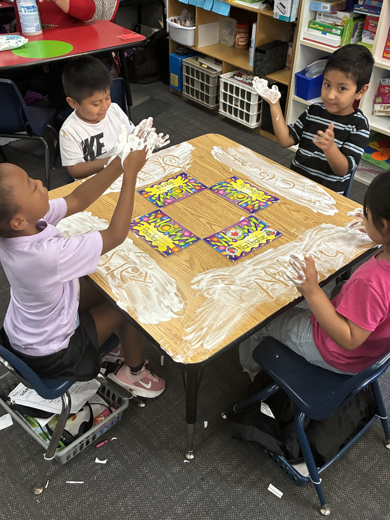 Four elementary students sit around a square wooden table in a classroom, engaged in a sensory learning activity. Each child has shaving cream spread in front of them with letters etched into it and raised, sudsy hands. Colorful name cards sit in the center of the table.