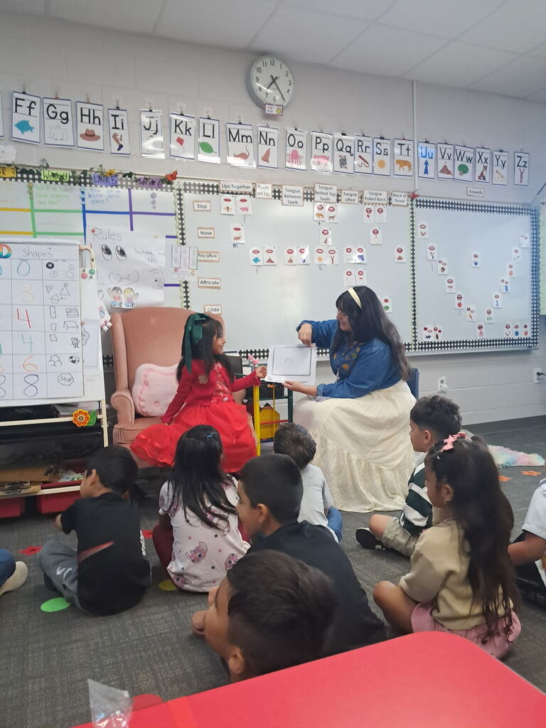A young girl in a red dress sits in a pink armchair while a teacher holds up a page of drawing to present to students sitting cross-legged on the rug.