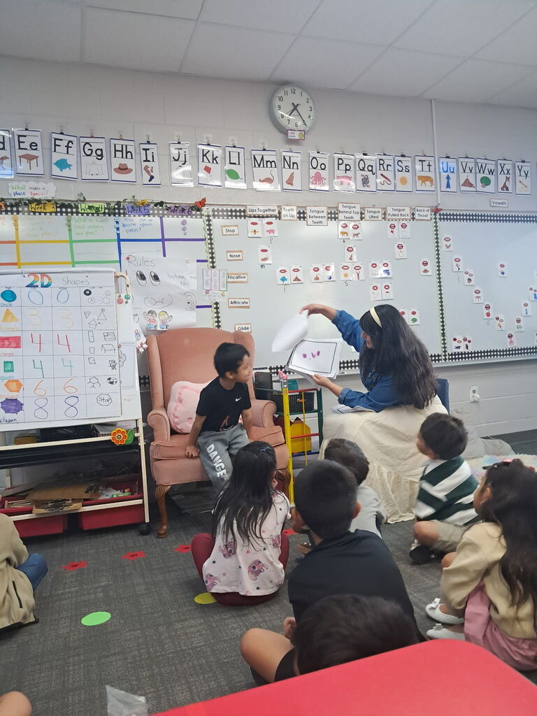A young boy stands next to a pink armchair while a teacher beside him turns pages on a display easel to showcase student artwork to the class.
