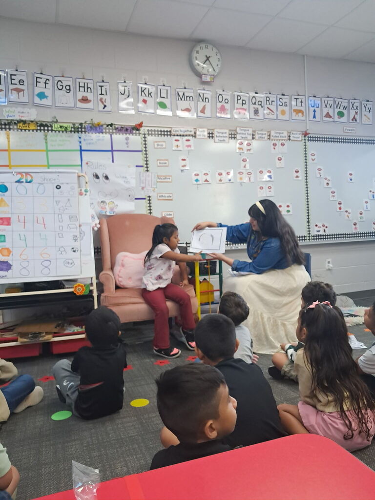 A young girl sitting in an armchair reaches out toward a picture being held by a teacher in a denim top and long skirt, while classmates watch from the floor.