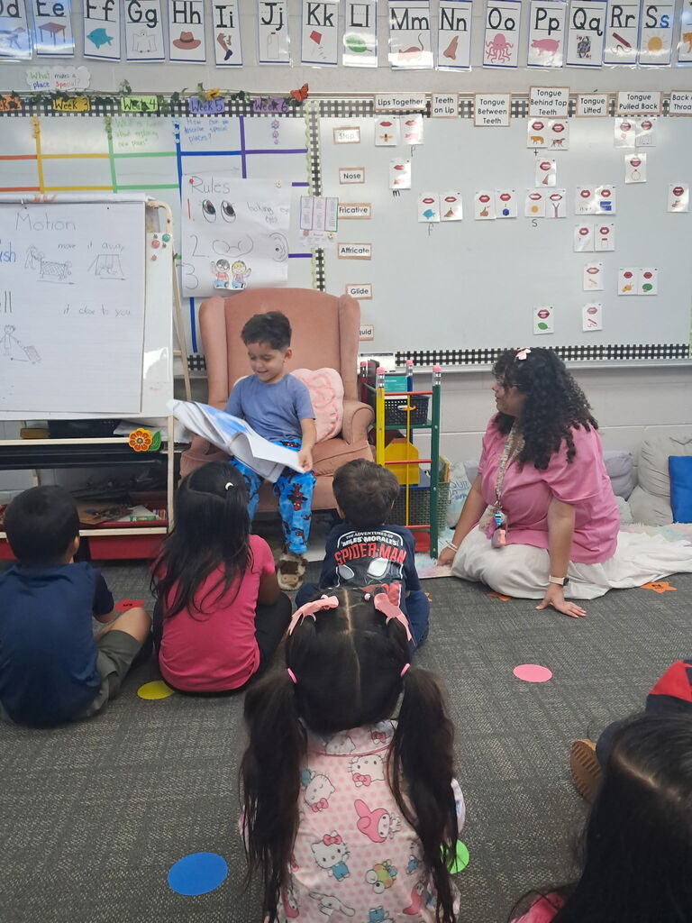A young boy in blue pajamas sits in a pink armchair reading a book aloud to his classmates on the carpet, as a teacher looks on supportively.
