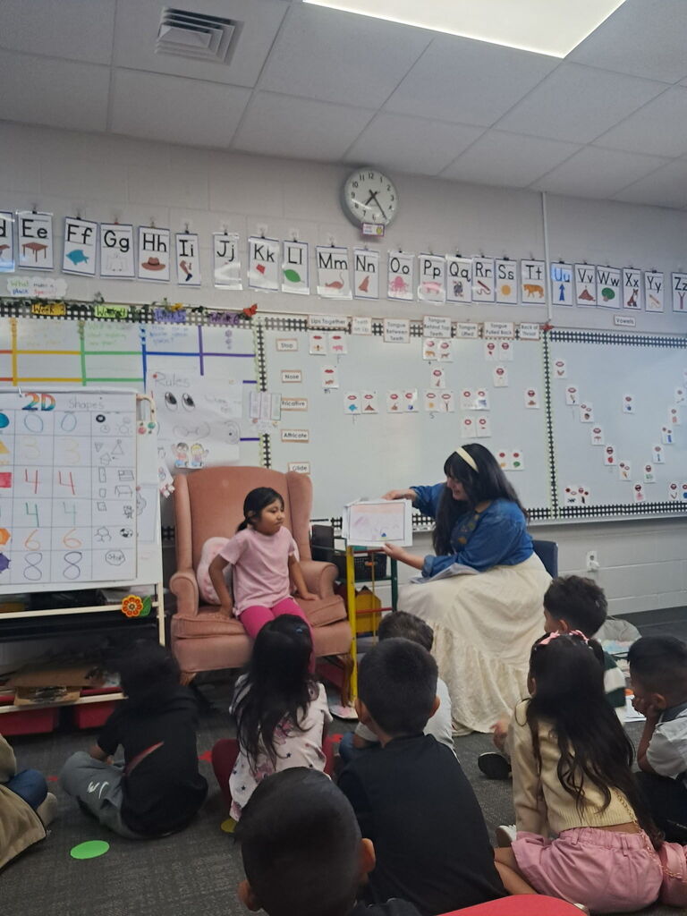 A young girl in a pink shirt sits in a pink armchair as a teacher next to her displays a student drawing to the class gathered on the carpet.