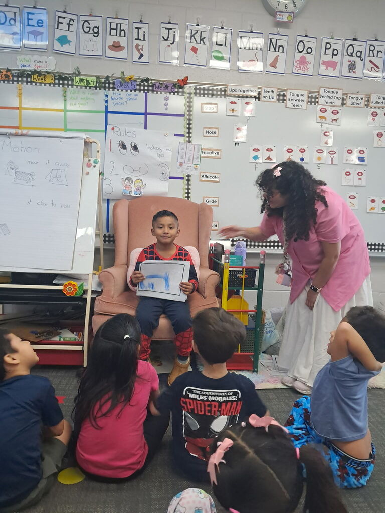 A young boy wearing a Spider-Man shirt sits in an armchair at the front of a classroom, proudly holding up a drawing to show his classmates on the carpet. A teacher stands beside him gesturing encouragement.
