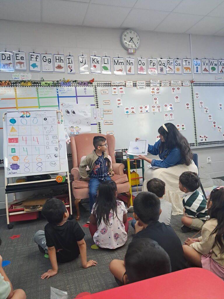 A young boy sits in an armchair as a teacher beside him holds up a paper drawing to present to students gathered on the rug in front of a white board.