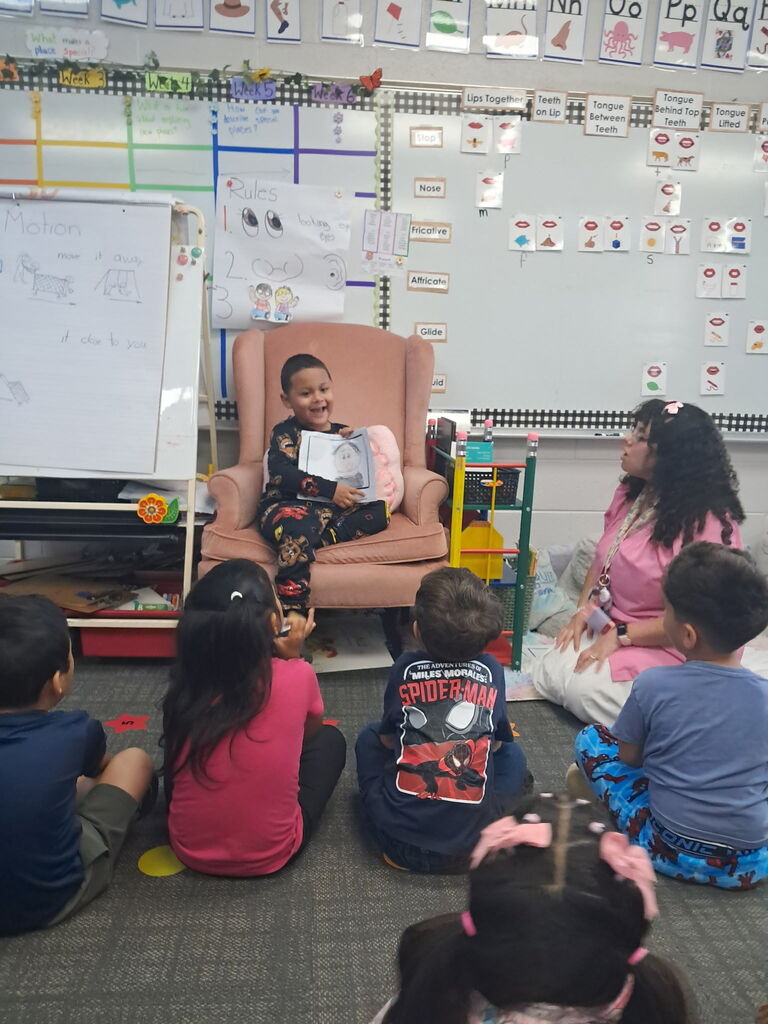 A smiling boy in dark pajamas sits in a pink armchair showing a drawing to his class, while a teacher in pink smiles at him from the side