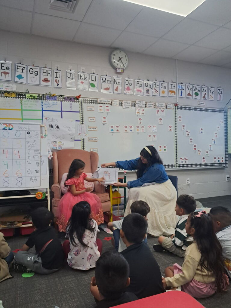 A young girl in a pink dress sits in an armchair while a teacher sitting next to her holds up a drawing for the class sitting on the rug to view