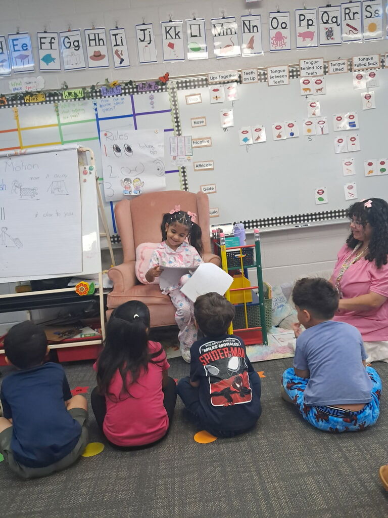 A young girl in pink pajamas sits in a large pink armchair in a decorated elementary classroom, holding papers and sharing a story with classmates seated on the rug. A teacher in a pink top sits nearby guiding the lesson.