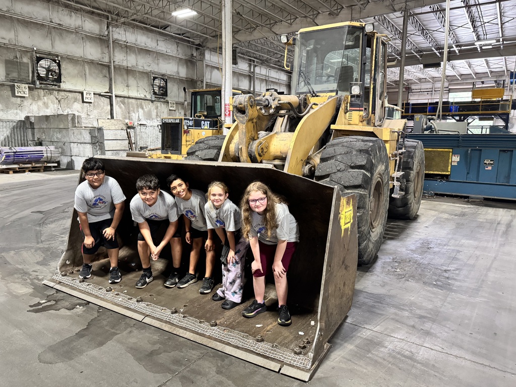 Five students wearing matching gray "Westwood School Recycling Team" t-shirts pose inside the large front bucket of a yellow wheel loader machinery inside an industrial recycling facility facility.