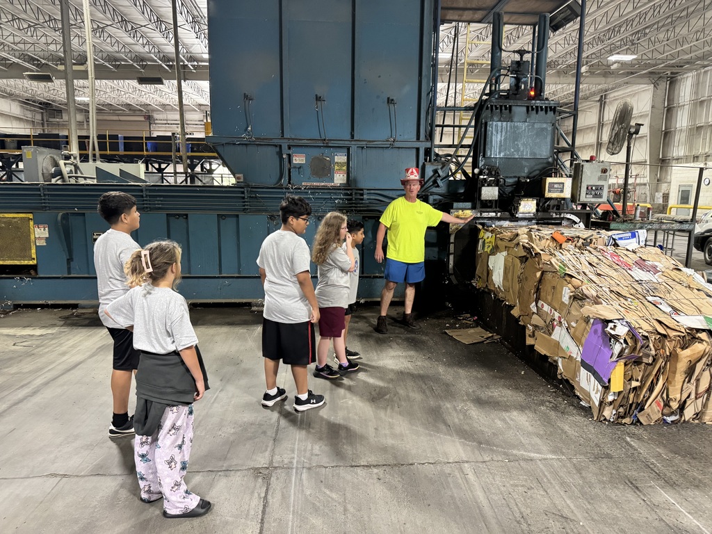 Students stand in a line inside a recycling facility, watching a guide in a bright neon shirt present a large, tightly compressed bale of crushed cardboard emerging from a industrial baler machine.