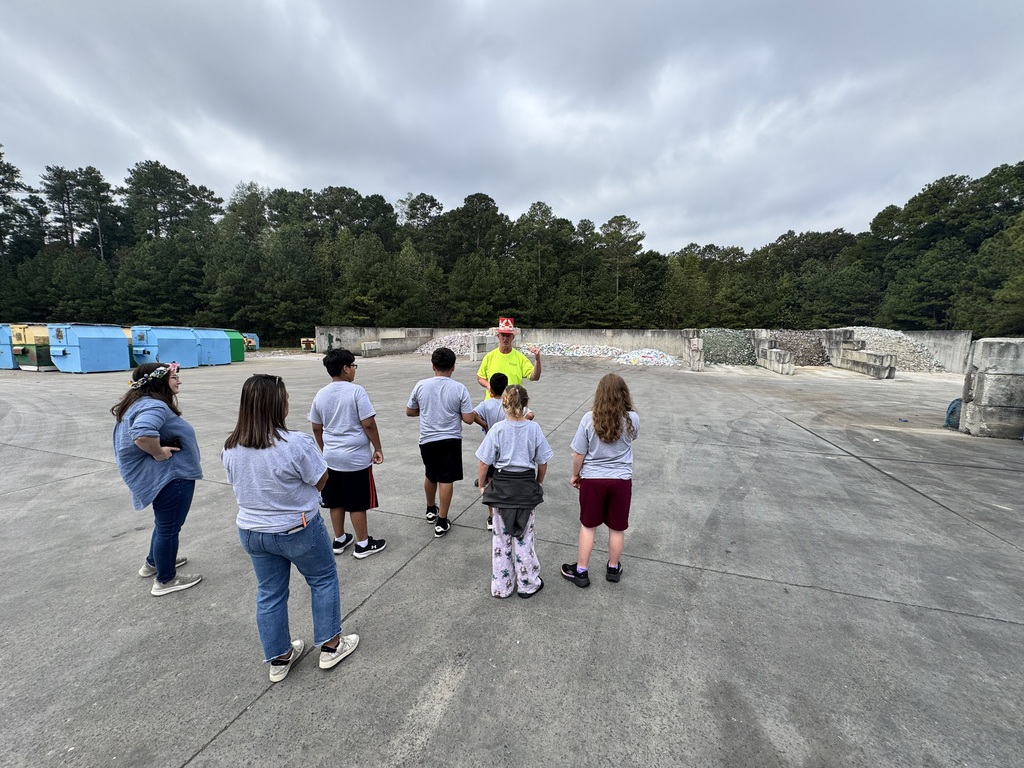 A group of students and adults stand in a semi-circle outdoors on a large concrete pad, listening to a guide in a bright neon shirt and hat. Large recycling collection containers and piles of sorted recyclable materials line the background perimeter under an overcast sky.