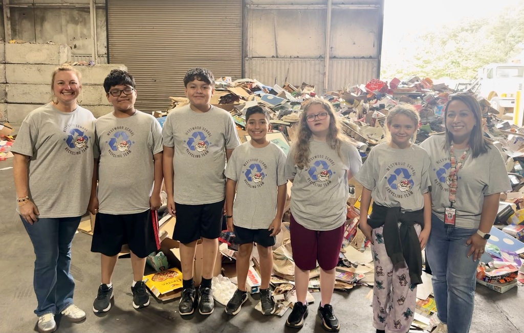 Students and adult chaperones, all wearing matching gray "Westwood School Recycling Team" t-shirts, line up together in front of a massive mound of recyclable cardboard and paper inside a processing facility.