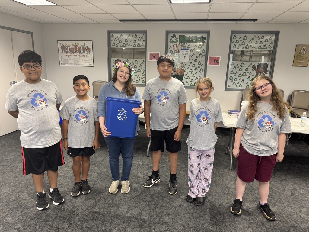 Five students wearing matching gray "Westwood School Recycling Team" t-shirts stand in a line alongside an adult wearing a floral crown, who holds a blue recycling bin in the center of the room. Recycling-themed posters line the wall behind them.