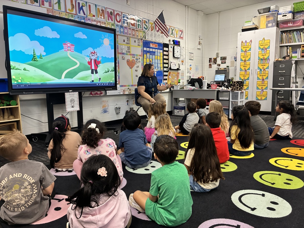 Elementary school students sit on a colorful smiley-face rug in a classroom, listening to a teacher read a picture book. Next to the teacher, a large interactive display screen shows a cartoon scene of a schoolhouse on a green hill with a mascot character.