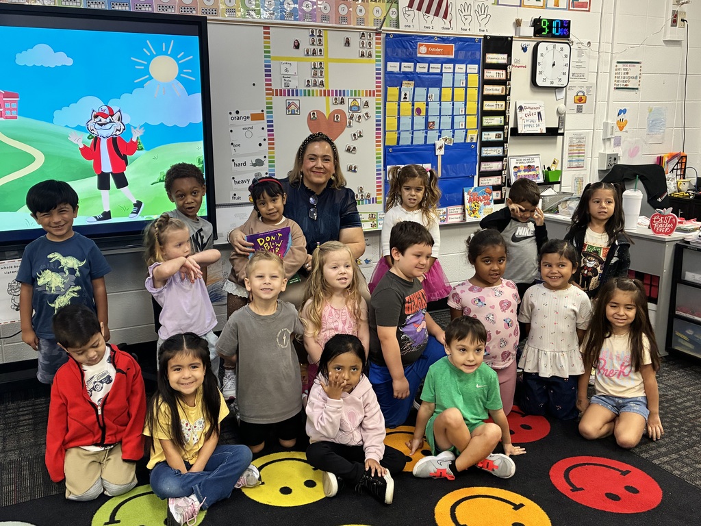 A group of young elementary students and their teacher gather together for a class group photo in a classroom setting. The teacher smiles from the center, holding a children's book, while students sit and kneel in front of her on a colorful smiley-face rug. A large interactive screen in the background displays a cartoon wildcat mascot against a sunny landscape.