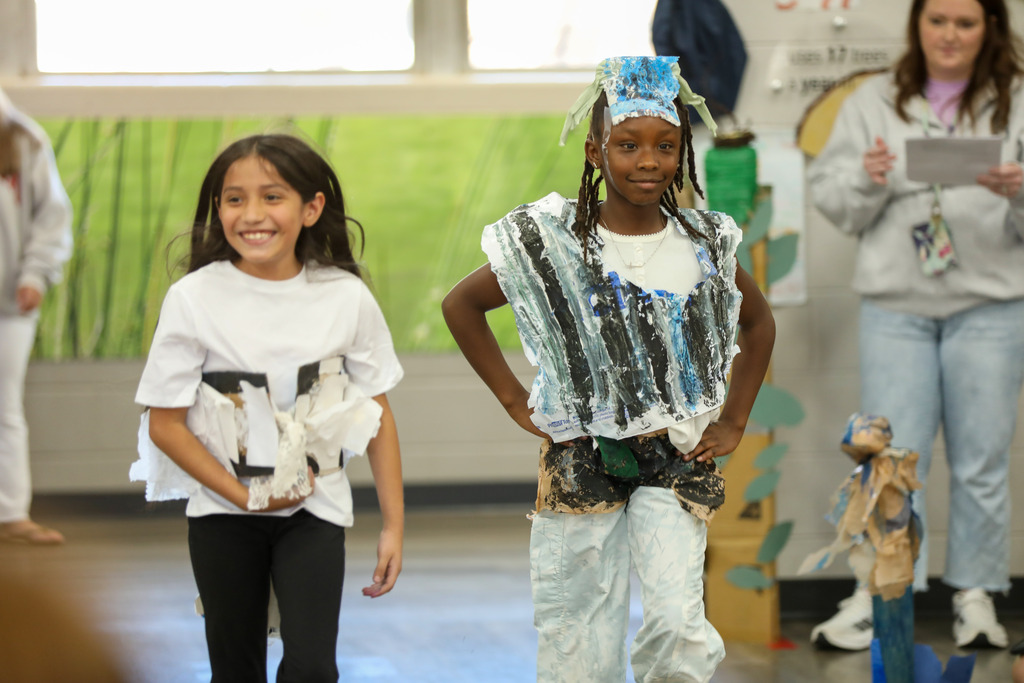 Two young students walk down a runway displaying handmade, recycled-material outfits during a school fashion show.