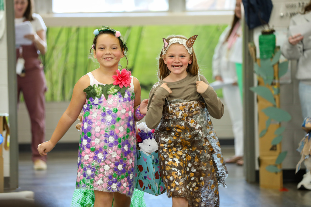 Two young girls walk down a runway wearing handmade, nature-themed dresses decorated with flowers and leaf patterns, smiling as they present their outfits.