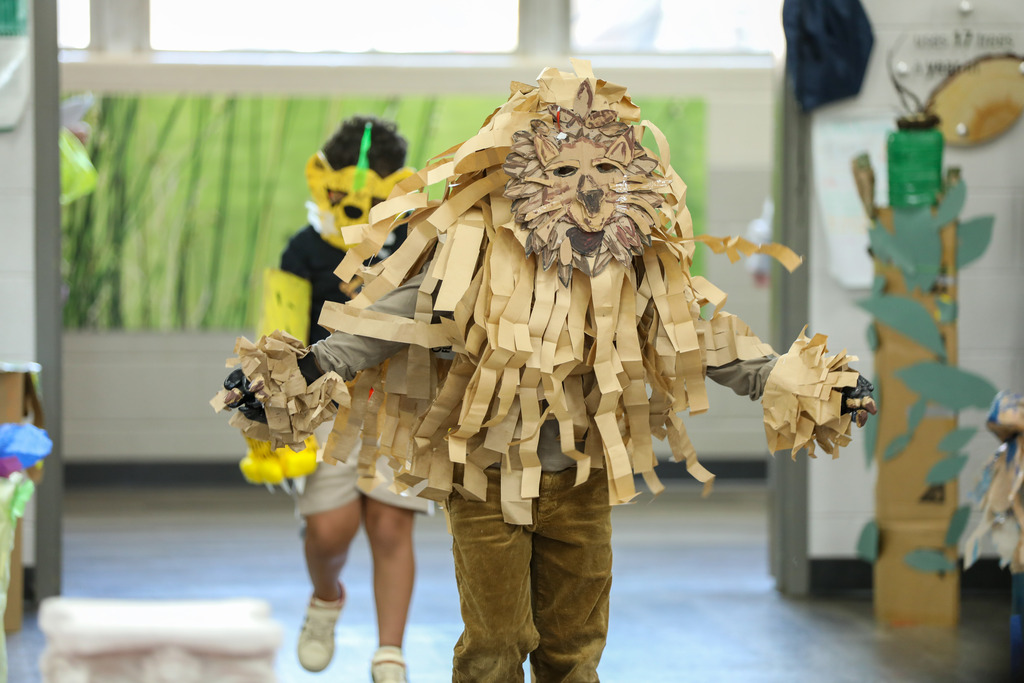 A young student wearing an elaborate, handmade lion costume made from shredded paper and cardboard walks down a pathway during a school event.