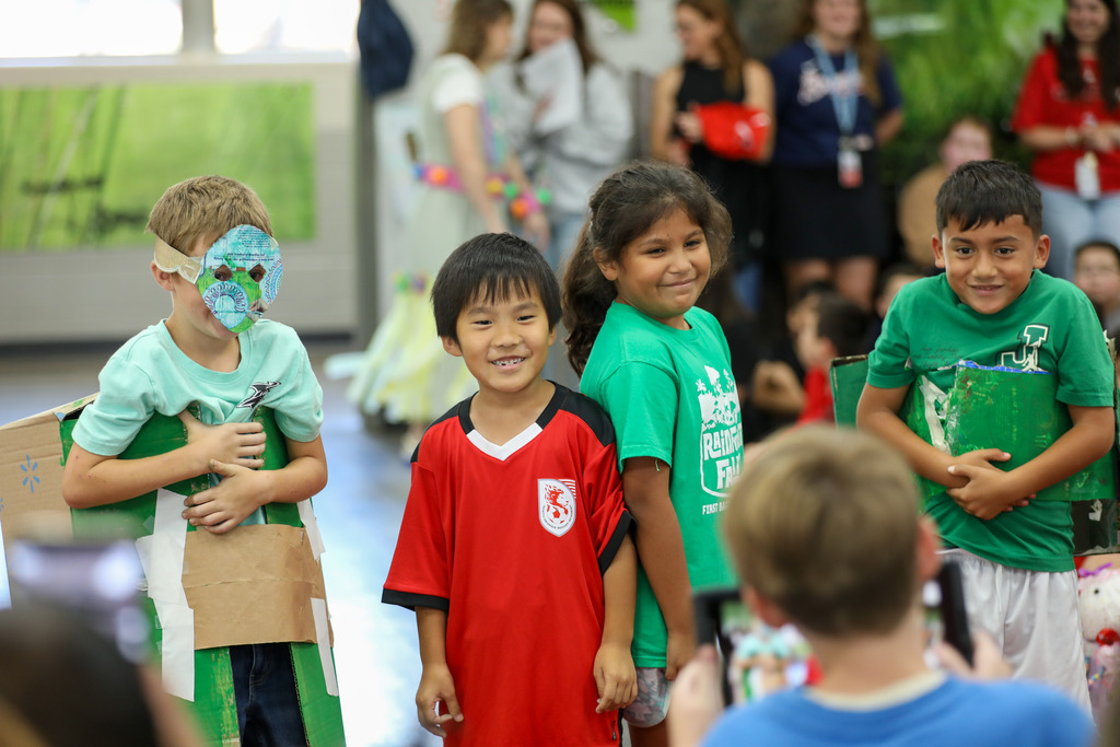 Young students wearing recycled-material costumes walk down a runway while a parent takes photos from the audience.