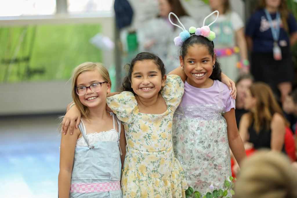 Three young schoolgirls wearing colorful dresses stand close together with their arms around each other, smiling at the camera.