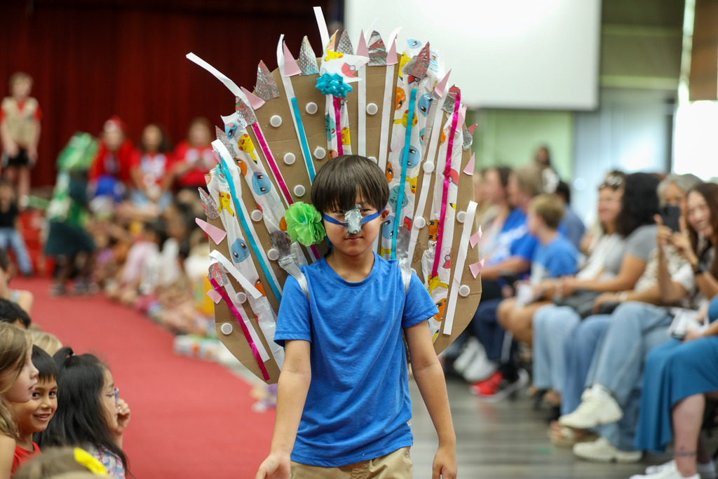 A young boy wearing a blue shirt and a mask walks down a carpeted runway showcasing a vibrant, winged costume made from cardboard, painted paper, and recycled plastic materials. Spectators sit on either side watching and taking photos.
