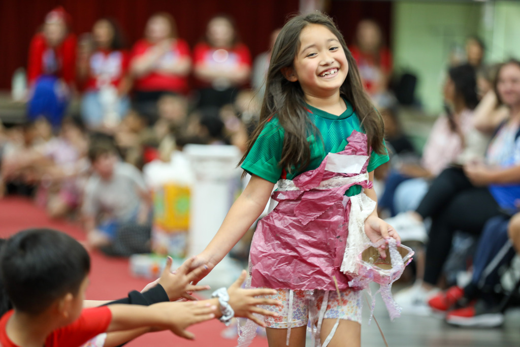 A smiling young girl walks down a runway wearing a handmade outfit fashioned from pink and green recycled plastic bags, high-fiving spectators in the audience.