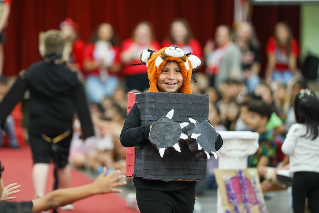 A young student wearing a handmade animal costume made from cardboard boxes and felt gloves walks down a red carpet runway in front of an audience.  