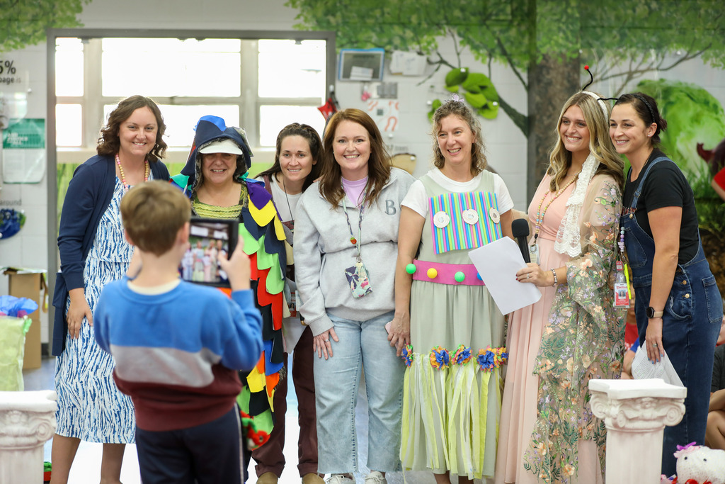 A group of six female teachers smiling together in front of a painted tree backdrop in a classroom. In the foreground, the back of a young student's head is visible as they participate in the school activity.