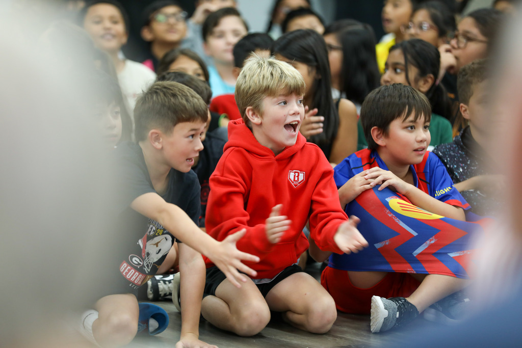 Young students sitting on the floor applaud and cheer for their classmates during a school fashion show.