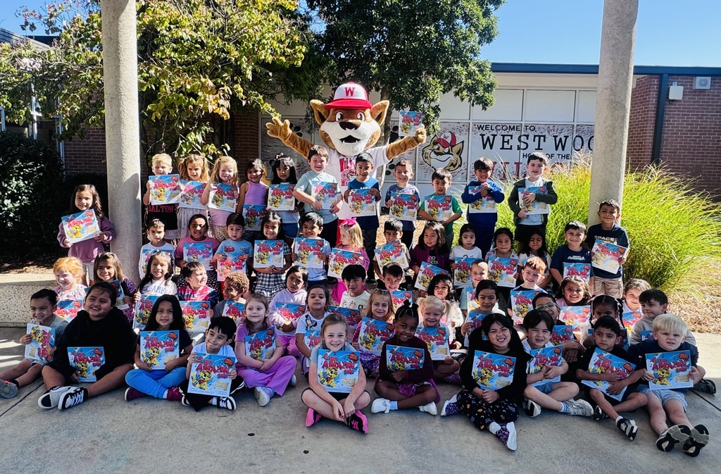 A large group of young elementary students poses outside in front of a school building with a sign reading "Welcome to Westwood." Standing behind the center of the crowd is a school mascot dressed as a wildcat wearing a white shirt and a red cap. Almost all of the children are sitting or standing while holding up copies of the same book, It’s an Amazing World!, showing off the cover.