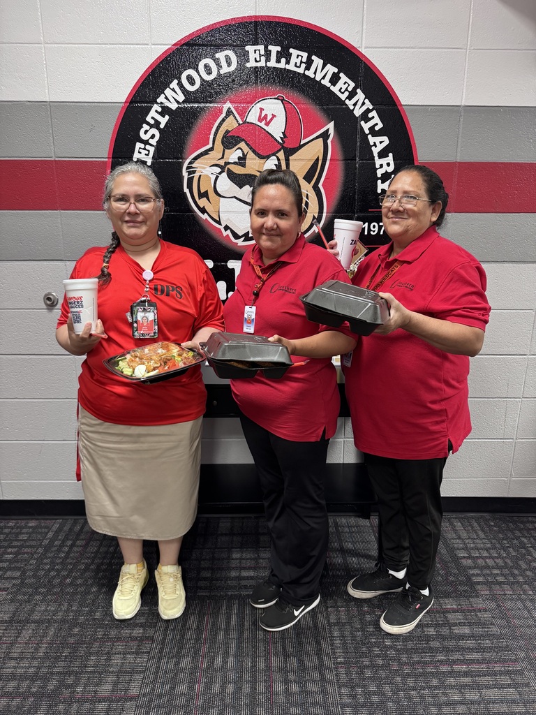 Three female staff members dressed in matching red polo shirts stand side by side in front of a painted circular Westwood Elementary logo featuring a wildcat mascot on a cinderblock wall. They are all smiling and posing with takeout food and drinks, including salads, black containers, and Zaxby's cups.