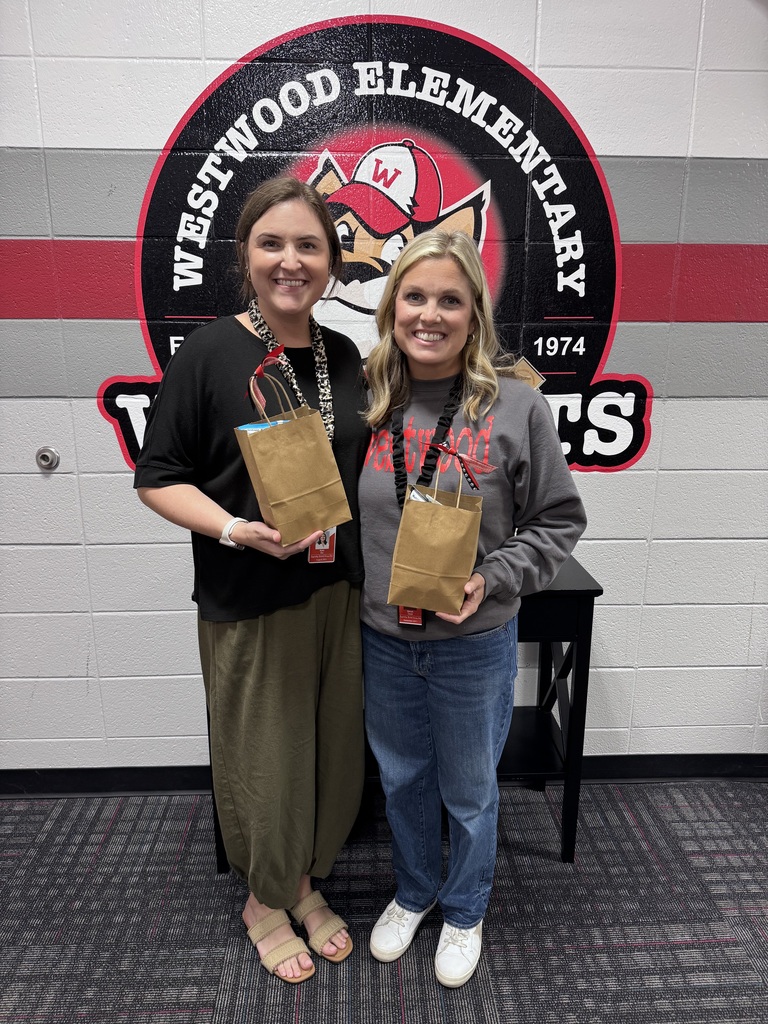 Two women stand together smiling in front of the Westwood Elementary wildcat logo painted on a cinderblock wall. The woman on the left wears a black shirt, olive green pants, and a leopard-print lanyard. The woman on the right wears a grey long-sleeve Westwood shirt, blue jeans, and white sneakers. Each holds a small brown paper gift bag tied with red ribbon.