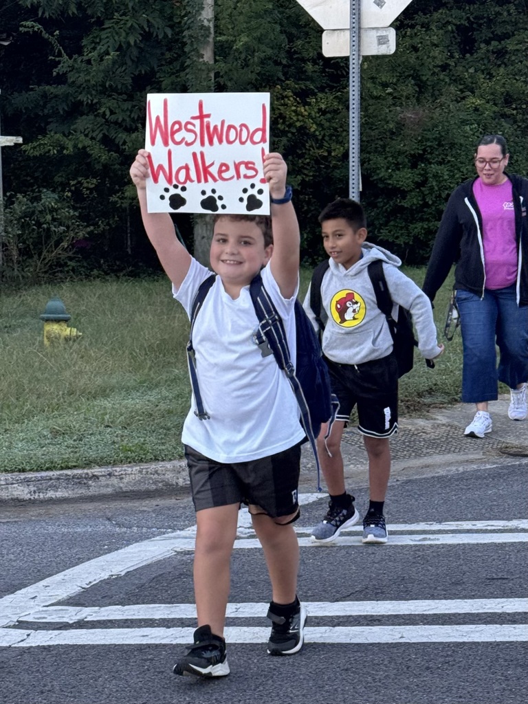 A young boy wearing a white t-shirt, dark shorts, and a backpack holds up a handwritten sign above his head that reads "Westwood Walkers!" in red lettering with black paw prints drawn below. He smiles as he steps through a painted crosswalk. Behind him, another student in a grey Buc-ee's hoodie walks alongside an adult in a pink shirt and black zip-up jacket.