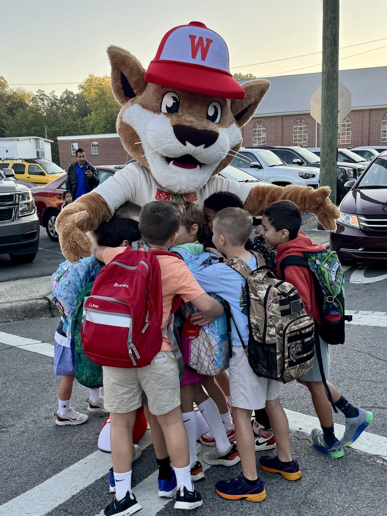 A wildcat mascot wearing a red baseball cap with a white letter "W" bends down to wrap its furry arms around a group of young school children wearing backpacks in a central group hug. The children surround the mascot cheerfully in a paved lot near parked vehicles.