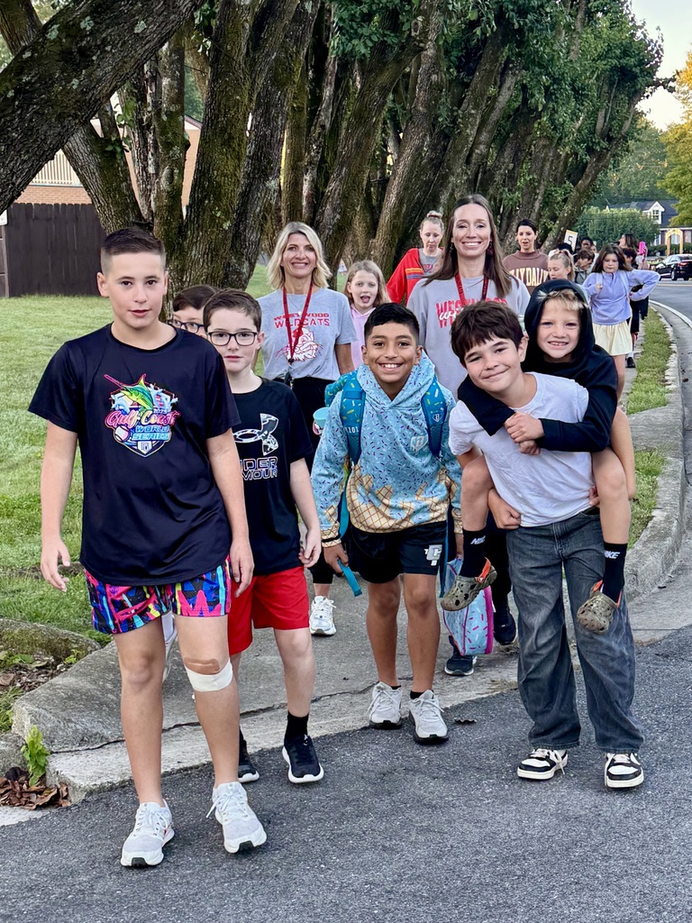 A cheerful group of elementary students walk along a curved, tree-lined sidewalk outdoors. On the right, a boy in a white t-shirt and grey jeans gives a piggyback ride to another boy wearing a black hooded sweatshirt. Next to them, a boy in a light blue patterned hoodie smiles at the camera. To their left walk two other boys—one wearing a black "Gulf Coast World Series" t-shirt with colourful patterned shorts and a bandage on his leg, and the other wearing a black Under Armour t-shirt with red shorts. Teachers and adult supervisors wearing Westwood shirts walk closely behind the group.