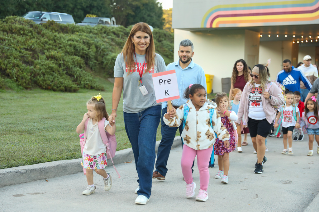 A smiling woman in a grey t-shirt and jeans holds hands with a young girl in a floral skirt and pink backpack as they walk along an outdoor paved path. Beside them, a young girl in pink pants and a teddy-bear pattern jacket holds up a sign that reads "Pre K". A man in a light blue shirt and other parents and children follow close behind them outside the school building.