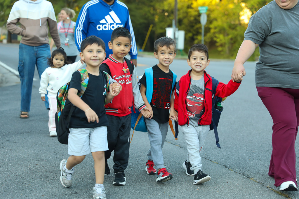 A group of four young school children wearing backpacks walk hand-in-hand down a paved road or parking lot. A toddler walks behind them, while adults accompany the group on either side. The children are smiling as they walk together outdoors.