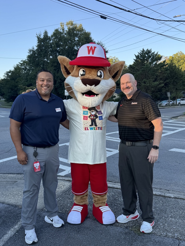 Two men in polo shirts stand smiling outdoors with their arms around a wildcat school mascot in the center. The mascot wears a red hat with a white "W" and a t-shirt featuring an illustration of the mascot in mariachi clothing with colorful papel picado flags and the text "El Westie". The group stands on a paved surface near a crosswalk and trees under a clear sky.
