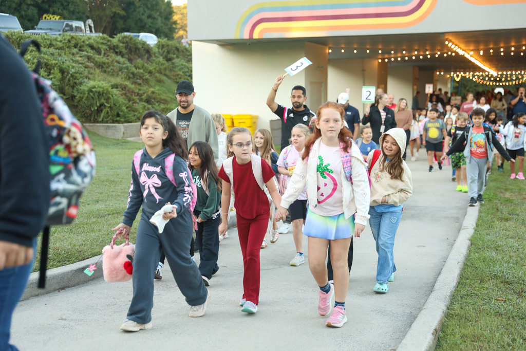 A group of elementary school students walk along a paved walkway outside a school building. In the front, two girls walk hand-in-hand: one in a red shirt and pants, and the other in a strawberry graphic shirt, light blue skirt, and white jacket. To their left, another girl in a dark grey tracksuit walks along carrying a pink plush handbag. Adults follow closely behind, with one man holding up a sign with the number "3" and another adult holding a number "2" sign further back.