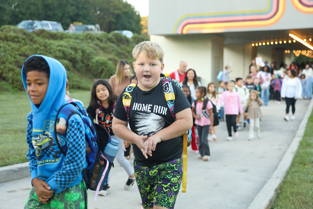 A young boy with blond hair wearing a black shirt with a "HOME RUN" baseball graphic and patterned shorts walks excitedly down a paved outdoor walkway. To his left, another boy walks forward wearing a bright blue hooded sweatshirt with a blue backpack. A large group of elementary students and accompanying adults stretch out in a line behind them outside the school.