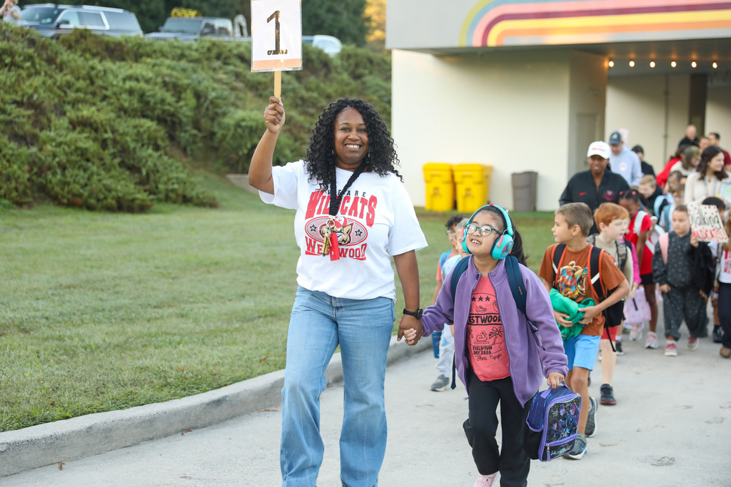 A smiling woman wearing a white "WE ARE WILDCATS" t-shirt and jeans leads a group of students outside. She holds up a wooden sign featuring the number "1" with one hand and holds the hand of a young girl with the other. The girl wears glasses, blue noise-canceling headphones, a purple jacket over a Westwood shirt, and carries a lunchbox. Other elementary students and adults walk closely behind them on the paved walkway.