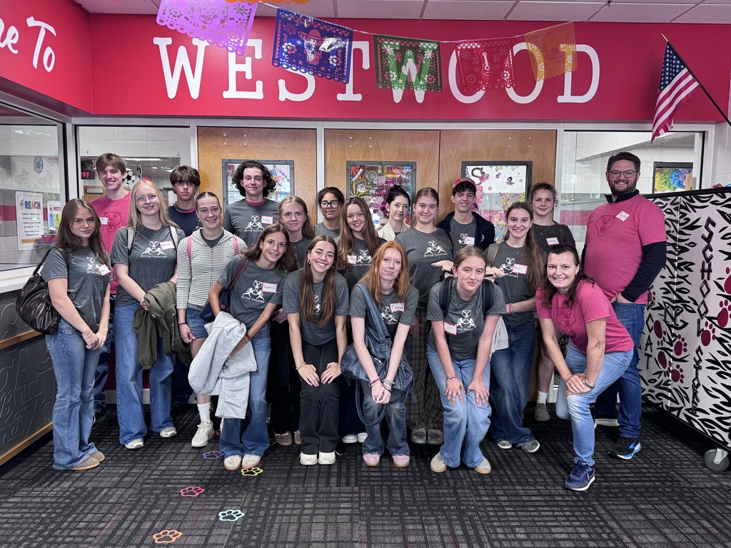 A group photo of about eighteen students and adults standing inside the lobby of Westwood School. A bright pink wall in the background features large white lettering that reads "WESTWOOD," colorful papel picado banners hanging above, and an American flag to the right. Most students in the group wear matching dark gray t-shirts featuring a white graphic logo with the text "Better Together," while a few adults and students wear bright pink shirts with a wildcat mascot logo.